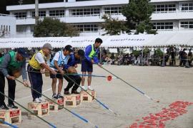 5名の参加者が、ビールケースの上に乗りながら、カギを使って、地面に沢山並べられている赤い貝のようなものを取ろうとしている写真