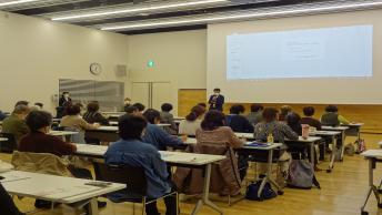 前方のスクリーンに写し出される資料をもとに講演をする男性と、多くの参加者の方々の写真