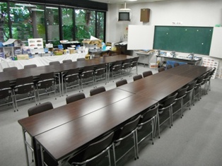 前に黒板があり、長テーブルにパイプ椅子が設置された集会室の写真