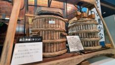 2つの大きな木製の樽に重しが乗っている左前方に粕圧搾機(キリン)の説明板がある様子の写真