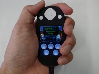 ひとつの縦長の画面と、複数のボタンのついた小さな楕円形の小型のタブレットの端末を、片手で握っている写真