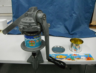 ハンドルがひとつついていて、上下から缶詰を挟める金属製の機械の真ん中に缶詰がひとつ置かれている写真