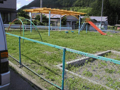 フェンスで囲われた芝生の中に雲梯、滑り台が設置されている、児童遊園の写真
