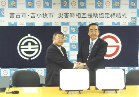 山本正徳宮古市長と岩倉博文苫小牧市長が両手で握手をしながら記念撮影をしている写真