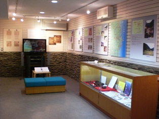 壁に数多く資料が展示され、その下に展示物が置かれたショーケースと牧庵鞭牛の資料用映像が流れそれを腰かけながら見ることができる椅子が置かれている展示室内の写真