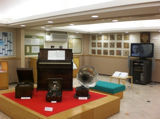 展示台の上に昔のオルガンや蓄音機などが展示され展示台の後方に鳥取春陽の資料用映像が流れそれを腰かけながら見ることができる展示室内の写真