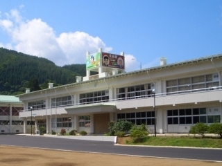 白の学校校舎に似た建物の上に、緑と赤の看板が立っていて、その看板には、人の顔が二人載っている新里分室(新里生涯学習センター 玄翁館内)の写真