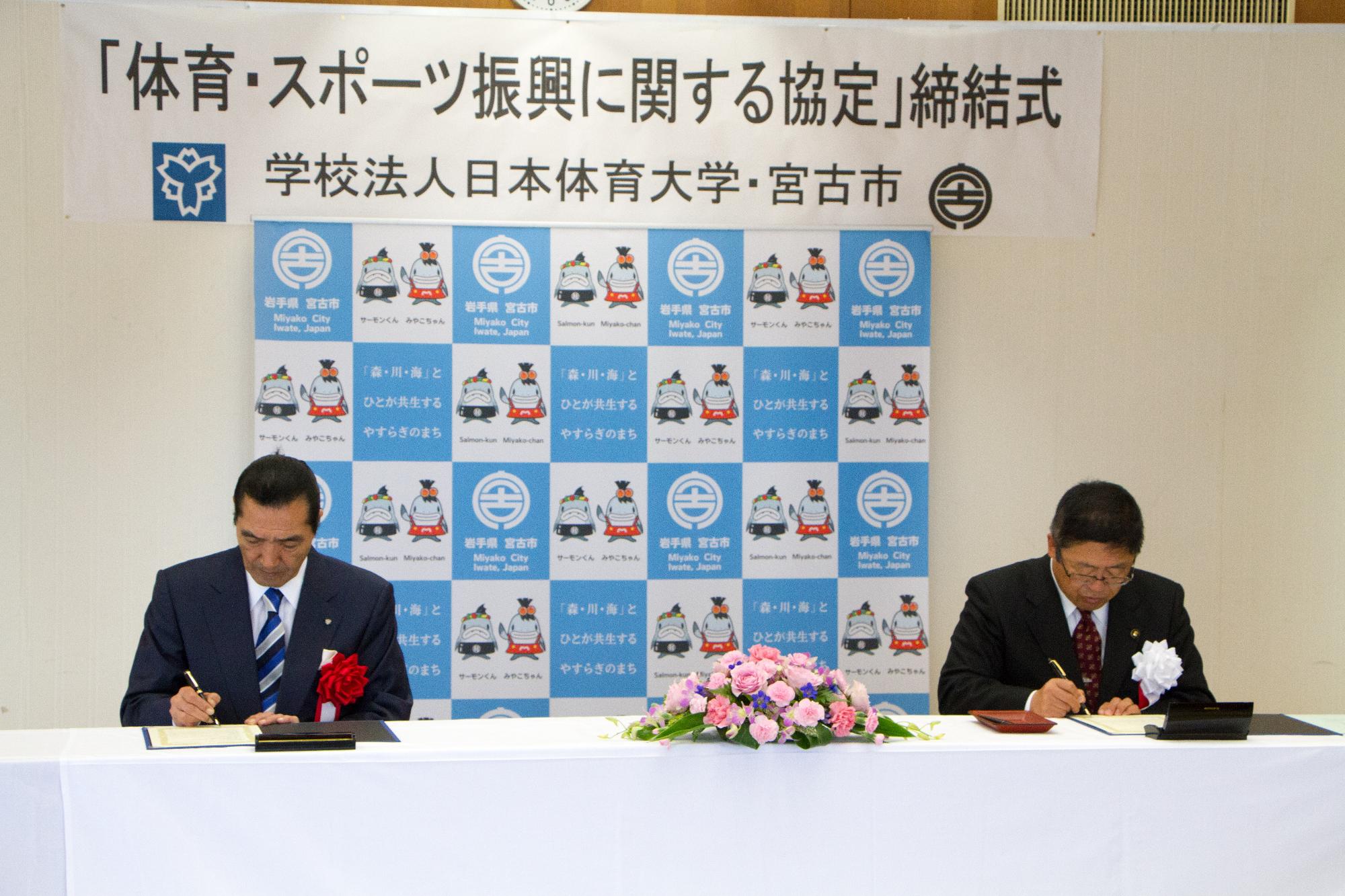 「体育・スポーツ振興に関する協定」締結式と書かれた横幕と宮古市のバックボードの前に白い布がかけられた長テーブルの前に松浪理事長と市長が座り、締結書にサインをしている様子の写真