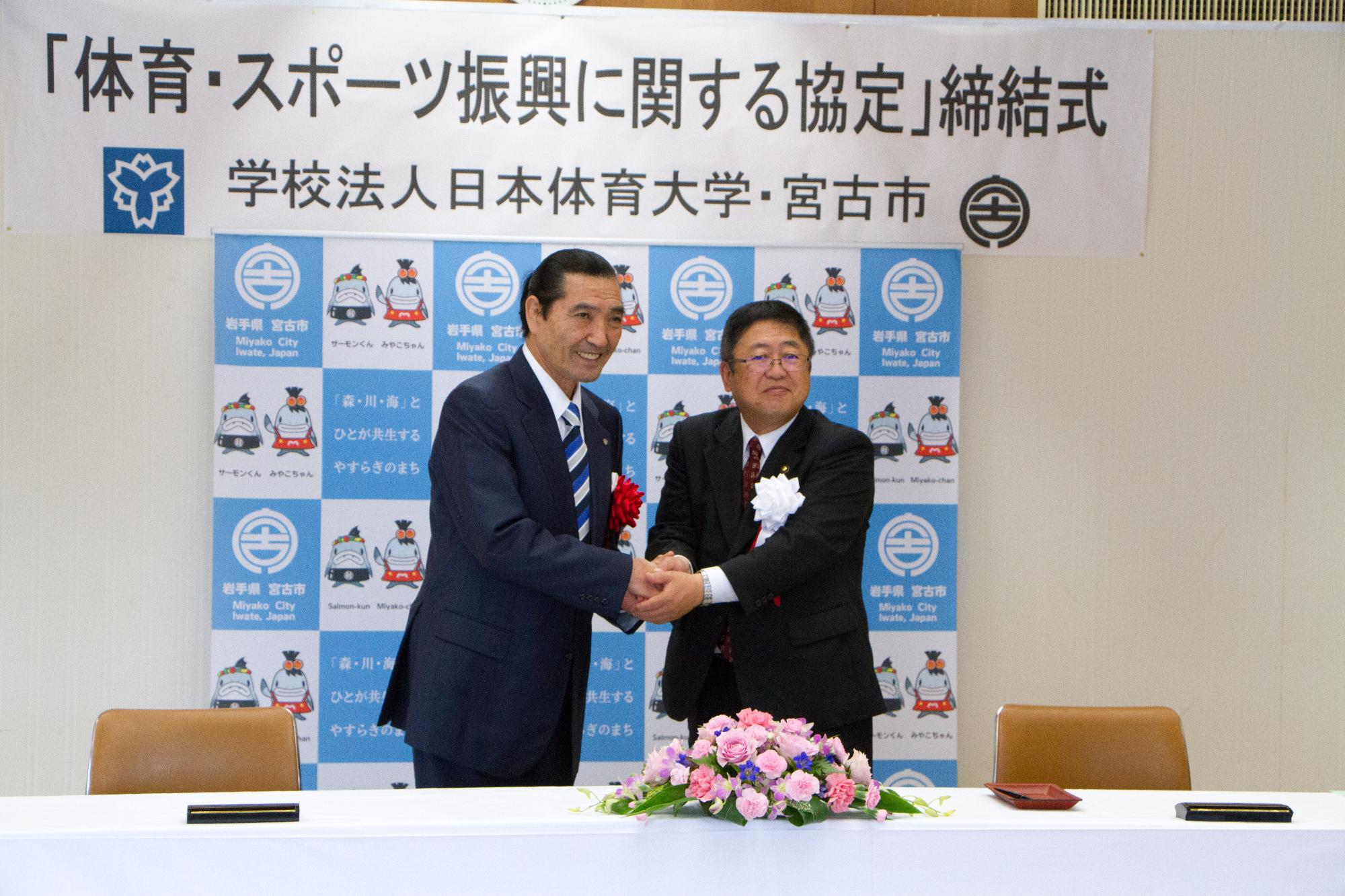 「体育・スポーツ振興に関する協定」締結式と書かれた横幕と宮古市のバックボードの前で松浪理事長と市長が両手で握手を交わしている写真