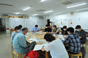 大きなテーブルに10人ほどが囲んで座り、資料などを見ながら話をしている様子の写真