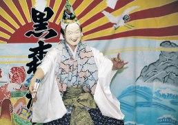 白地に青や赤の刺繍が施され金色の袴を着用し能に使うお面を付けた演者が太陽や鶴、魚などの縁起が良いものが描かれた大きな幕の前で扇子を持って夜神楽をしている写真