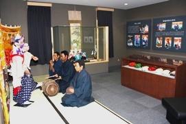 展示室内の一角で二枚の畳を敷き、白い衣装を纏った人の前に男性三人が正座をし 太鼓や笛などで演奏をしている写真