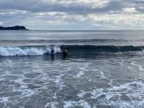 モクモクとした雲が浮かび薄っすらと太陽の光がさしてきた空の下、打ち寄せる波に乗りサーフィンを楽しんでいる親子の写真