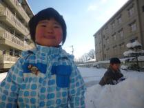 団地に挟まれた広場で、雪に埋もれて遊ぶ男の子や左手前で水色のジャンパーを着て真っ赤な頬っぺたをした子どもがカメラ目線で笑顔で立っている写真