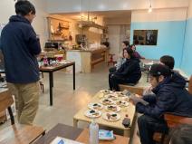 ゲストハウス内の大きなテーブルや低いテーブルの上に料理が並び、左側に立っている男性の話を椅子に座っている参加者が聞いているおためしツアーの様子の写真