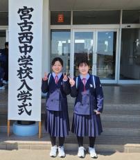 校舎の入り口に「宮古西中学校入学式」と書かれた看板が立つ傍で、真新しい紺色の制服を着用した2名の女子生徒がカメラに向かってピースサインをしている写真