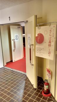 白色の壁に1と書かれ、扉にも1と書かれた張り紙が貼られた会場の入り口の右側に、「「さらば義経」を語る」と書かれたポスターがポールに掲示されている写真