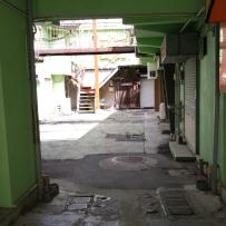 緑色の外壁が建つ路地の奥に光が差し込んだ広場とお店が建っている路地裏の写真