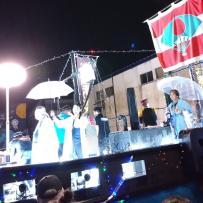 雨が降っているステージ上で傘をさしながら披露している演者たちの写真