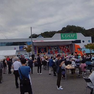 大漁旗が飾られたステージの前に椅子に座ったり立って眺めている来場者が大勢いる産業まつりの様子の写真