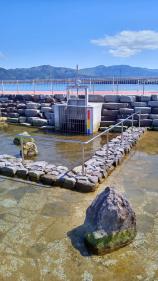 遠くに山並みや海が見える公園内にある、石で囲まれ手すりが設置されている潮だまりの写真