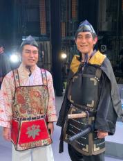 右側の男性は2本の刀を差して黒色の甲冑を着用し、左側の男性は薄ピンク色の着物に花柄模様の甲冑を着用した市民劇「さらば義経」に参加した2名の男性が笑顔で並んでいる写真