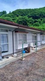 木々が生い茂る手前の敷地内に赤い屋根の平屋建ての住宅があり、窓にはレースのカーテン、庭には物干し台が設置されている田舎体験住宅の建物外観の写真