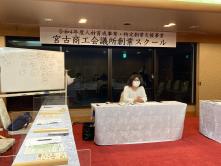 宮古商工会議所創業スクールと書かれた横断幕が設置された赤色の絨毯の会議室に白色のテーブルクロスが掛けられた長机が設置されており、奥の席に座った女性講師による講義が行われている様子の写真
