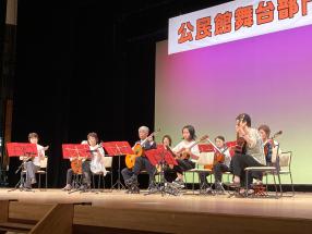 ステージ前列にギターを持った5名と後列にマンドリンを持った3名のメンバーが椅子に座り、演奏を行っている公民館舞台部門発表会の様子の写真