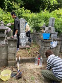 草木に囲まれたお墓の前にバケツや箒が置かれ、若い男性や女性が墓掃除を行っている様子の写真