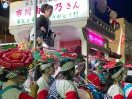 赤い牡丹や蓮華をかたどった造花を付けた妻折笠を被った踊り手が踊っている傍で、山車の上に乗り観客に手を振る黒と白の着物を着た市川由紀乃さんを撮影した写真