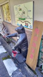 近隣の地図が壁に設置された駅舎沿いに立つ「熊出没注意」と書かれた看板と、手に赤色のペンキが入ったバケツを持って立っているクマの置物の写真