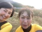 太陽の光がさしている海岸をバックに、黄色と黒色のウエットスーツを着用した笑顔の2名の女性がアップで自撮りで撮影した写真