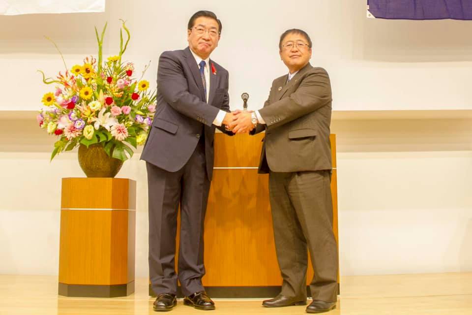 演台の前で、両手で固い握手を交わす老松大仙市長と山本宮古市長の写真
