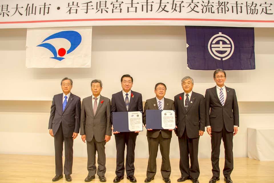協定書の横断幕や市章の前で、佐藤大仙市副市長、金谷大仙市議長、協定書を持った老松大仙市長、協定書を持った山本宮古市長、古舘議長、佐藤副市長が横一列に並んでいる集合写真