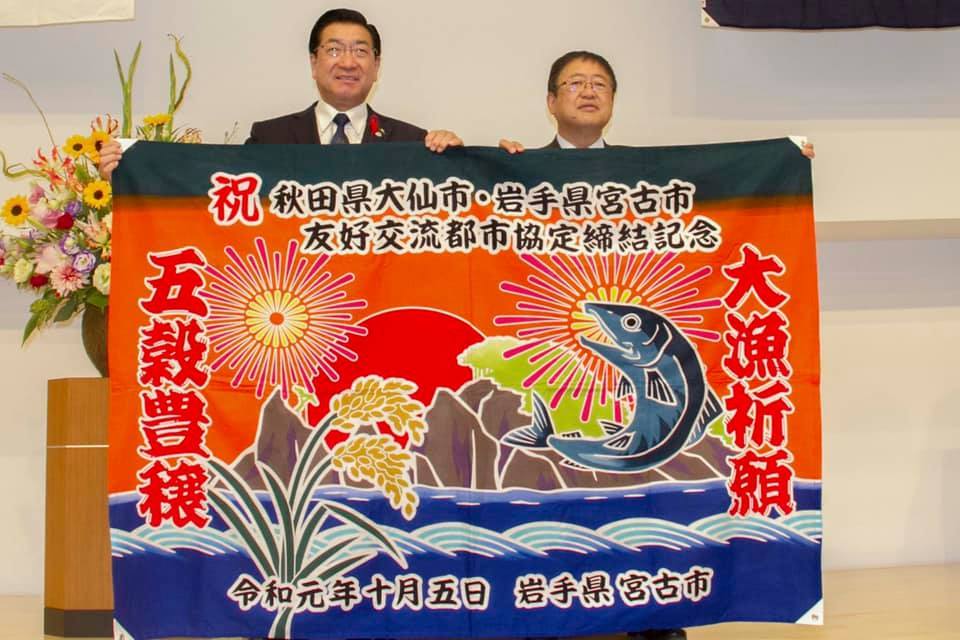 祝秋田県大仙市・岩手県宮古市友好交流都市協定締結記念と書かれた花火や鮭の絵が描かれた大漁旗を、老松大仙市長と山本市長が両手で広げ持っている様子の写真