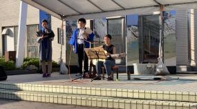 建物前に簡易のステージを設置し、3名がギターなどの演奏をおこなっている写真