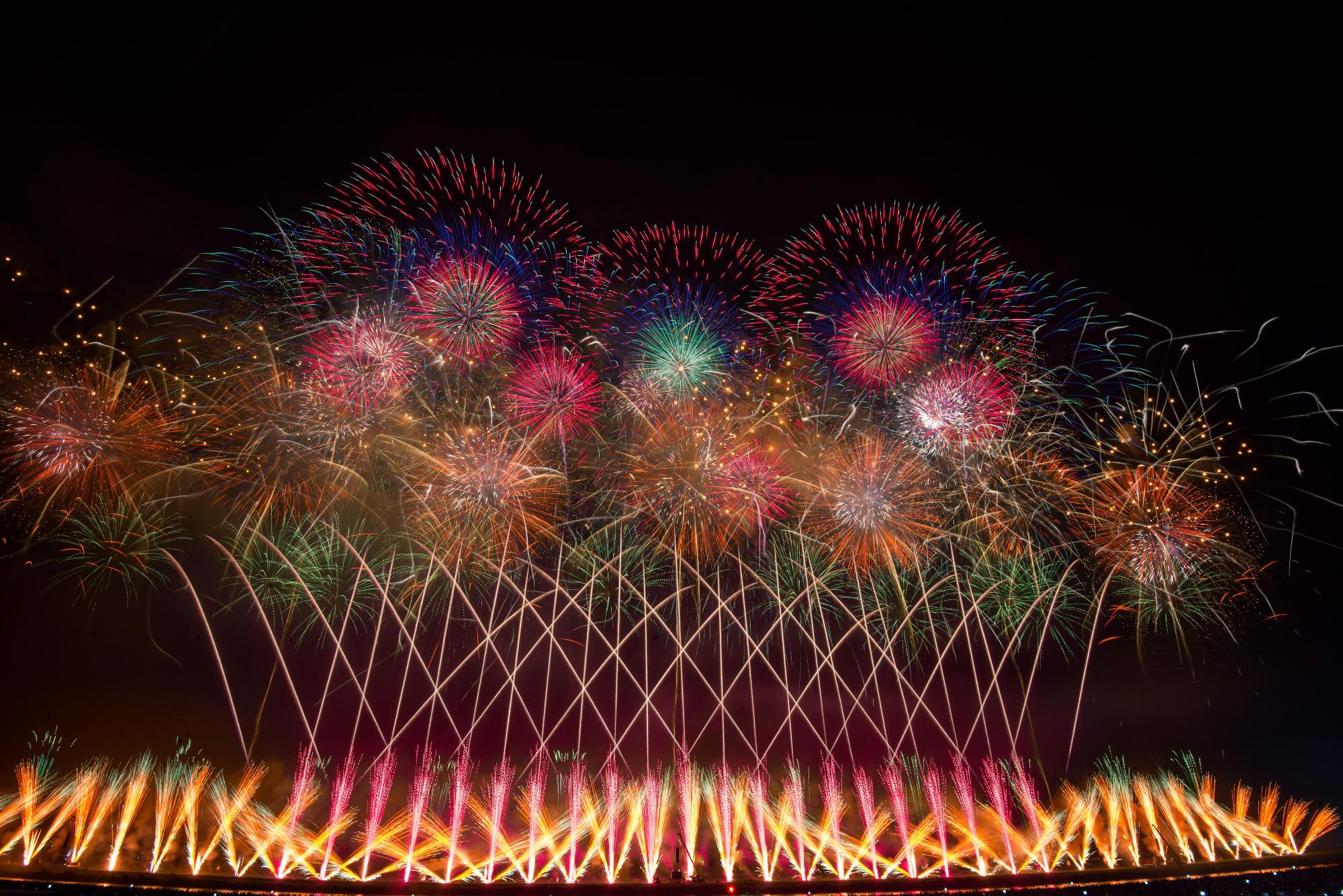 夜空に色とりどりの大輪の打ち上げ花火が打ち上がり、その下から複数の花火が交差になり夜空に上がっている花火の写真