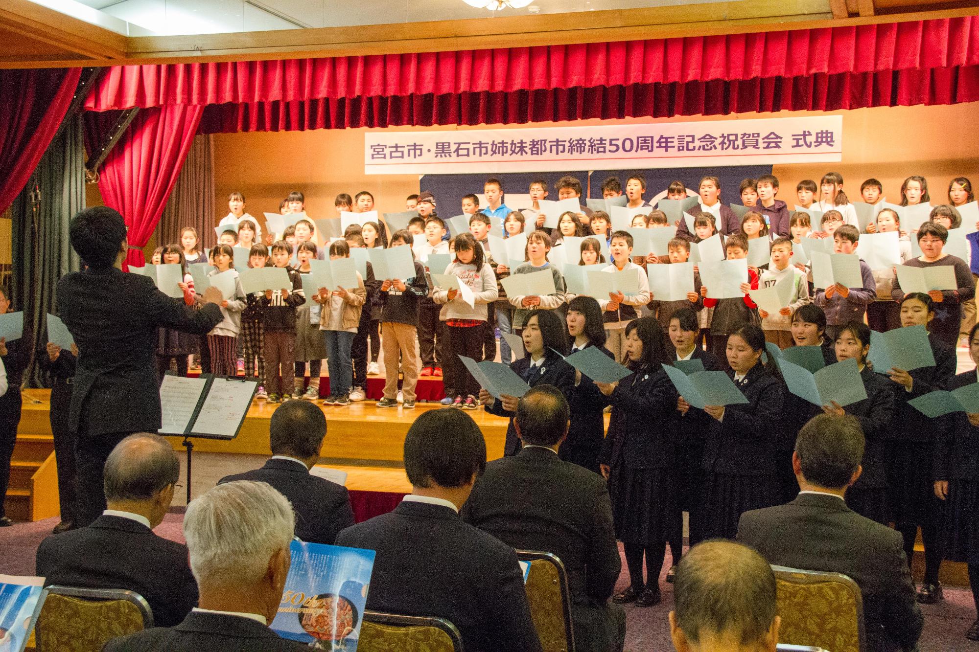 宮古市・黒石市姉妹都市締結50周年記念祝賀会式典と書かれた舞台に児童が並び、舞台下に制服姿の生徒が楽譜を持って歌ってる写真
