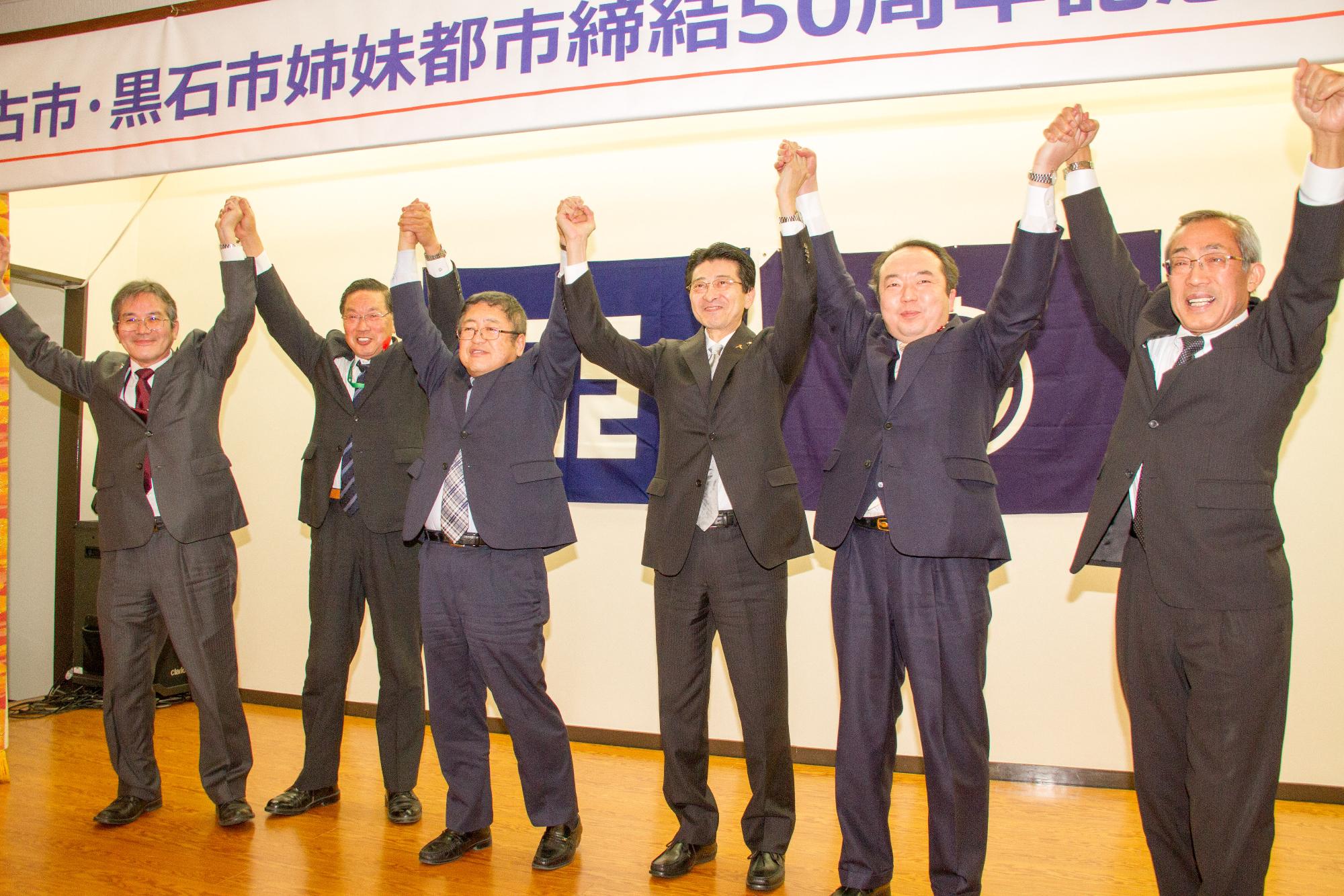 宮古市・黒石市姉妹都市締結50周年記念祝賀会式典と書かれた舞台でスーツ姿の6人の男性が横一列になり手を繋ぎ上げている集合写真