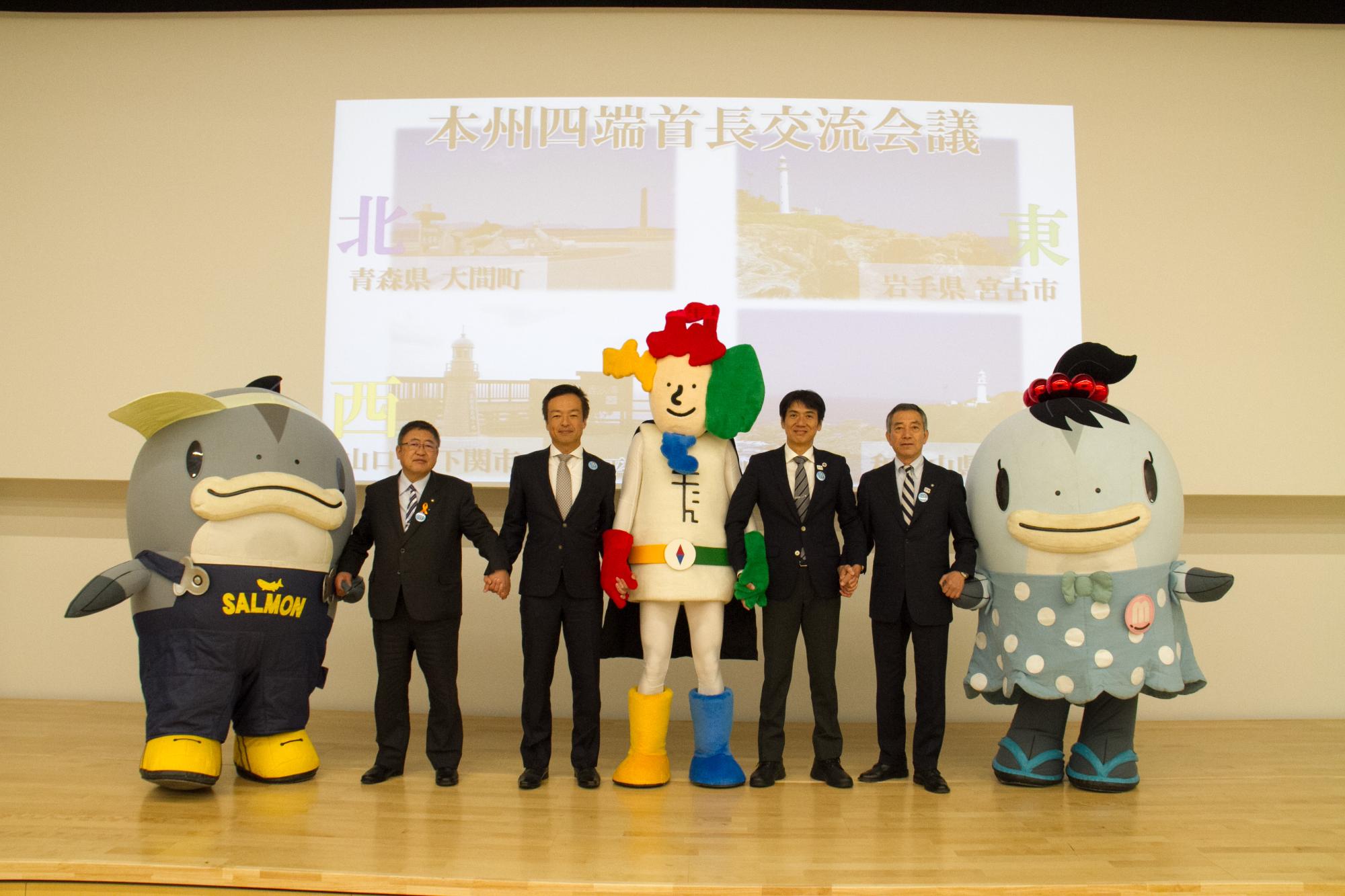 2018首長交流会議が行われたステージ後方のスクリーンに「本州四端首長交流会議」と書かれ4市の端っこの写真が映し出され、本州四端のヒーロー「よんたん」を中央に山口県下関市、和歌山県串本町、青森県大間町、宮古市の首長と宮古市のマスコットキャラクター「サーモンくん」と「みやこちゃん」が手をつないで横一列に並んでいる写真
