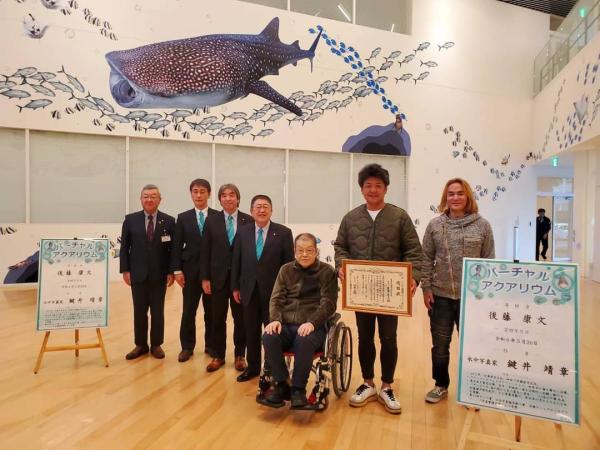 大きなジンベエザメや魚の大群が描かれたイーストピアみやこ館内の白い壁や柱の前で、賞状を両手で持つ鍵井靖章さんと車椅子に座る寄贈者の後藤 康文さんと山本市長や関係者合計7人の集合写真