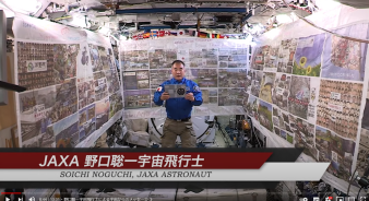 宇宙ステーション内で復興10年を物語る写真を刷り込んだ横断幕に囲まれて、メッセージを読み上げているJAXAの宇宙飛行士の写真