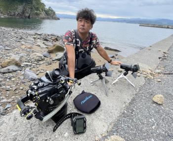 穏やかな海を背景に、岩がゴロゴロとした浅瀬でカメラの道具を広げて見せる水中写真家の鍵井 靖章さんの写真