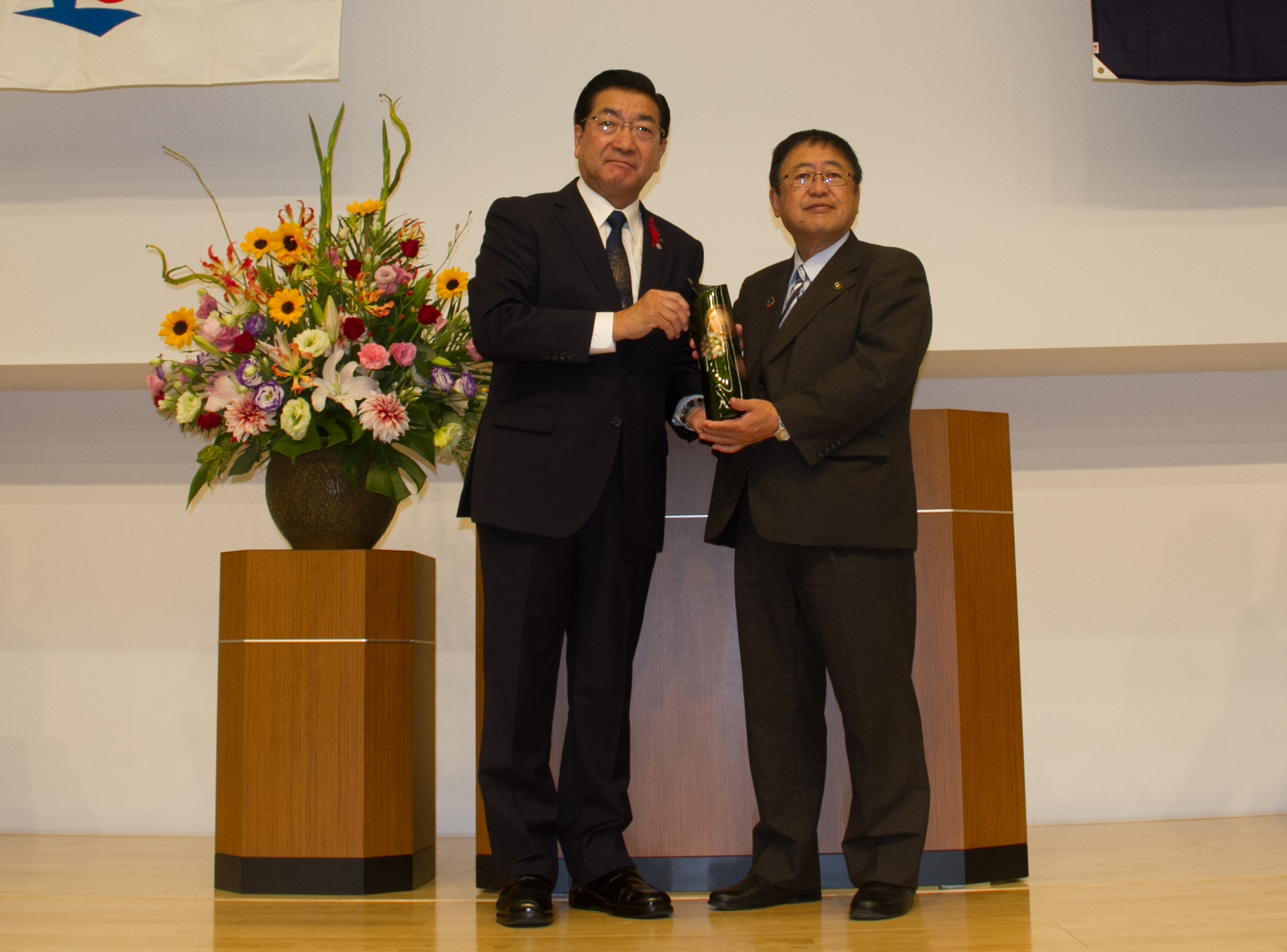 演台の前で、大曲の花火をイメージした川連塗りの花瓶を両手で持つ、老松大仙市長と山本宮古市長の写真