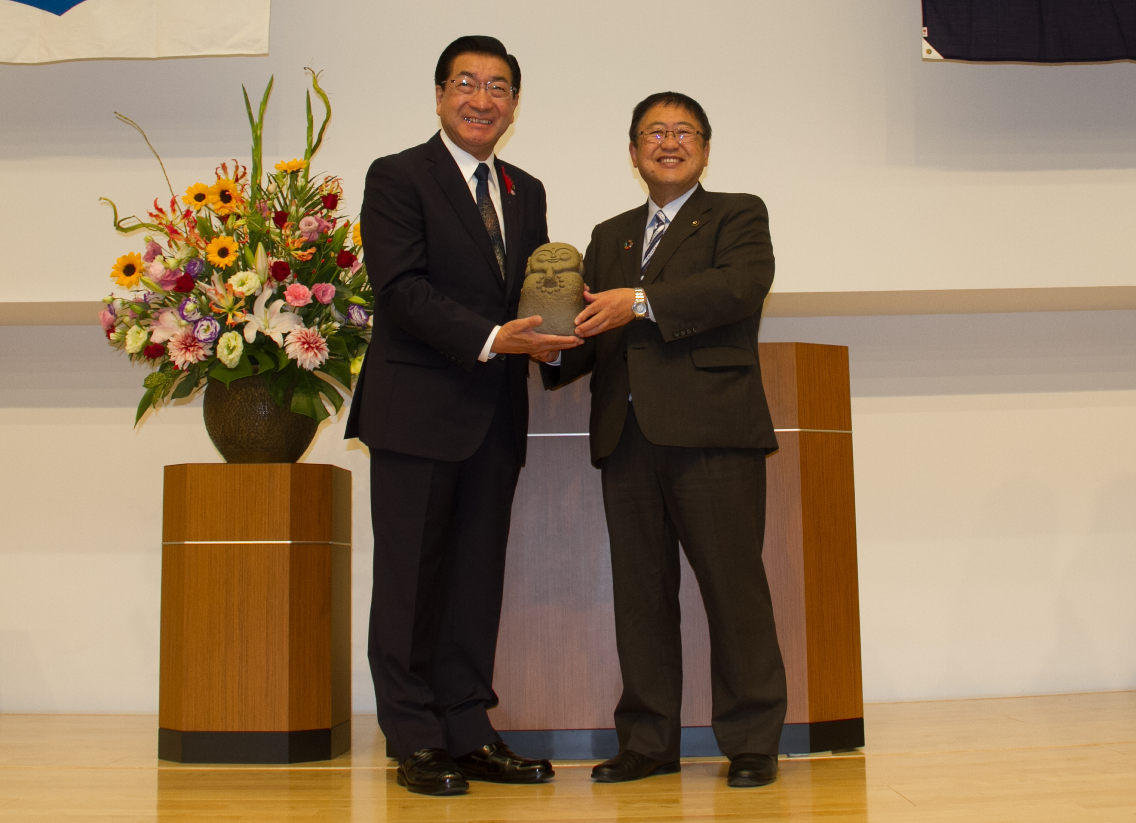 檀上に設置された演台の前で、1体のやすらぎ地蔵を老松大仙市長と山本市長が両手で抱えながら、笑顔で記念撮影に応じている様子の写真