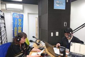 二人の男女がFM826と書かれた張り紙がしてある部屋で、マイクの前でラジオの収録をしている写真