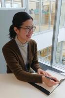 テーブルにノートを置いて片手にペンを持ちながら笑う女性を、斜め前から写した写真