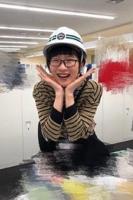 ヘルメットをかぶりながら、両手をあごの下に当て頬杖をつくようなポーズをとって笑う女性を正面から写した写真