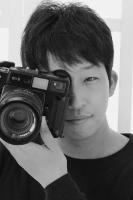 ほほえみながら片目をカメラのファインダーに当てて写真を撮る様子の男性を正面から写した写真